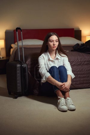 Foto de Triste joven mujer se sienta en el suelo cerca de una cama grande y una maleta junto a ella en la habitación. Check-in hotel - Imagen libre de derechos