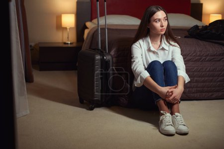 Foto de Hermosa joven sentada en el suelo cerca de una cama grande y una maleta junto a ella en la habitación. Check-in hotel - Imagen libre de derechos