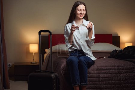 Foto de Mujer sonriente sosteniendo un vaso de agua y una tableta mientras está sentada en una cama grande en la habitación de hotel - Imagen libre de derechos