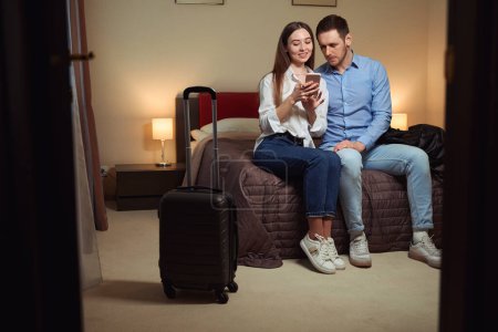 Téléchargez les photos : Couple amoureux naviguent ensemble sur le téléphone tout en étant assis sur un grand lit dans la chambre. Arrivée hôtel - en image libre de droit