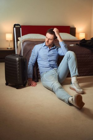 Foto de Hombre cansado sosteniendo un vaso de agua y una tableta mientras está sentado en el suelo cerca de la cama en la habitación del hotel - Imagen libre de derechos
