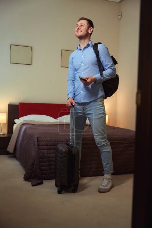 Téléchargez les photos : Beau jeune homme avec barbe tenant sac à dos et bagages debout dans la chambre. Check-in hôtel - en image libre de droit
