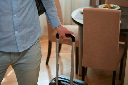 Téléchargez les photos : Jeune homme tenant la poignée de sa valise debout dans une chambre d'hôtel. Photo recadrée - en image libre de droit