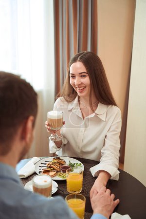 Foto de Una pareja joven y cariñosa desayunando sentada a la mesa en un hotel luminoso. Check-in hotel - Imagen libre de derechos