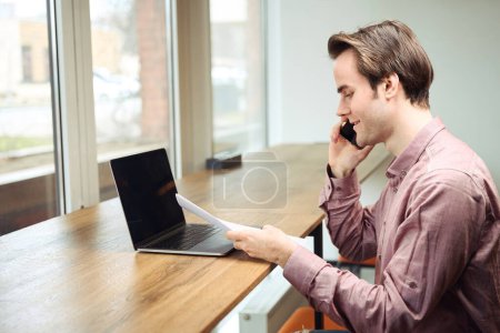 Foto de Vista lateral del empleado de la oficina sonriente sentado en el portátil mirando el documento en la mano mientras habla en el teléfono inteligente - Imagen libre de derechos
