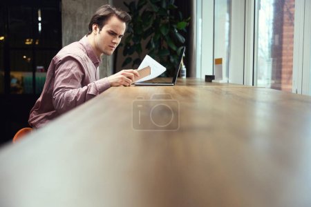 Téléchargez les photos : Homme avec un document à la main fixant l'écran du smartphone tout en étant assis à l'ordinateur portable dans un café - en image libre de droit