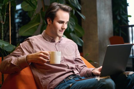 Photo for Pleased young man seated in armchair with coffee mug in hand typing on laptop keyboard - Royalty Free Image
