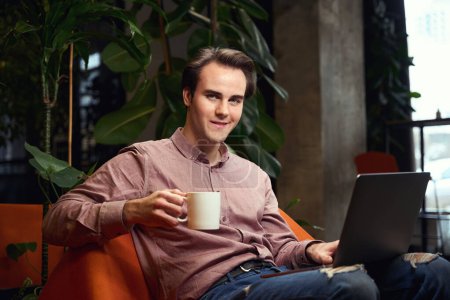 Téléchargez les photos : Jeune homme souriant avec tasse en céramique à la main et ordinateur portable assis dans un fauteuil dans un espace de coworking - en image libre de droit