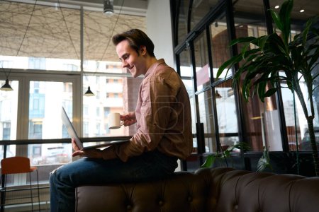 Photo for Side view of joyous freelancer with laptop and cup in hands sitting on sofa arm - Royalty Free Image