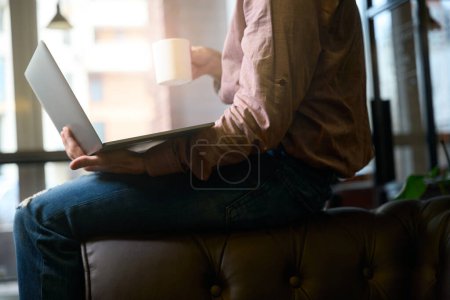 Téléchargez les photos : Photo recadrée de l'homme avec ordinateur portable et tasse dans les mains assis sur le canapé dans l'espace de coworking - en image libre de droit