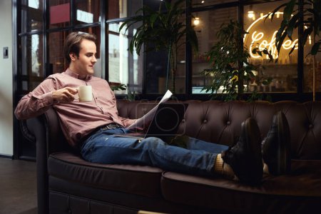 Photo for Pleased freelancer sitting on sofa with portable computer and pile of documents in hand during coffee break - Royalty Free Image