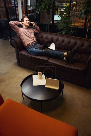 Téléchargez les photos : Jeune homme souriant avec ordinateur portable et smartphone à la main assis sur un canapé dans un espace de coworking - en image libre de droit