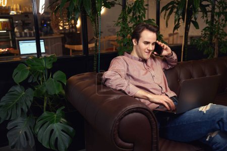 Photo for Joyful guy seated on sofa talking on smartphone while typing on laptop keyboard - Royalty Free Image
