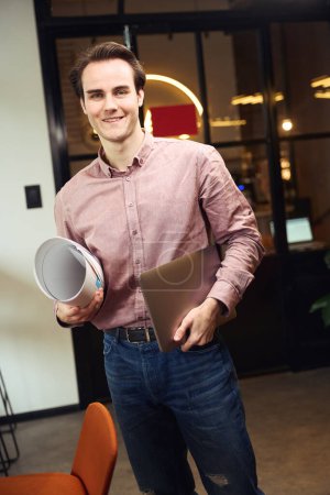 Téléchargez les photos : Jeune homme souriant avec ordinateur portable et dessin technique enroulé debout dans un espace de coworking - en image libre de droit