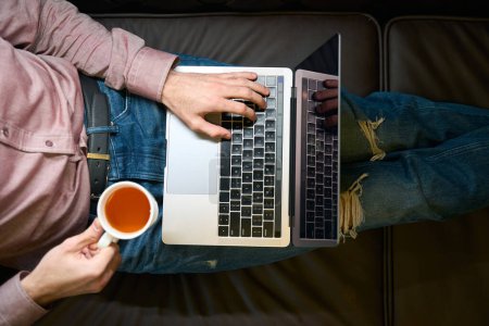 Téléchargez les photos : Vue du dessus de l'homme assis sur le canapé avec tasse de thé tapant sur le clavier de l'ordinateur portable - en image libre de droit