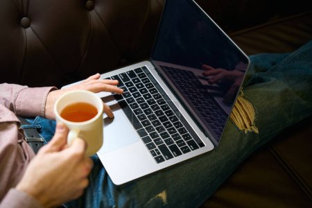 Téléchargez les photos : Photo recadrée d'un homme assis sur un canapé avec une tasse de boisson à base de plantes en cliquant sur l'index sur le pavé tactile de l'ordinateur portable - en image libre de droit