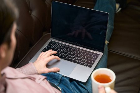 Téléchargez les photos : Vue arrière du mâle assis sur le canapé avec boisson à base de plantes en cliquant sur le pavé tactile de l'ordinateur portable - en image libre de droit