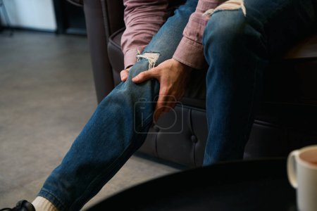 Foto de Foto recortada del hombre tocando su rótula mientras está sentado en el sofá de cuero - Imagen libre de derechos