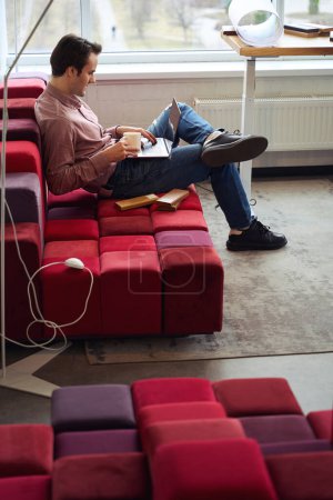 Téléchargez les photos : Vue latérale d'un professionnel du design calme et concentré assis sur un canapé avec des échantillons de bois travaillant sur un ordinateur portable dans son bureau - en image libre de droit