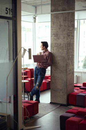 Téléchargez les photos : Travailleur d'entreprise calme avec ordinateur portable dans les mains appuyé contre pilier dans le bureau pendant la pause café - en image libre de droit