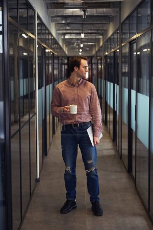 Foto de Retrato de larga duración del trabajador corporativo tranquilo con portátil y taza en las manos de pie en el pasillo de la oficina vacía - Imagen libre de derechos