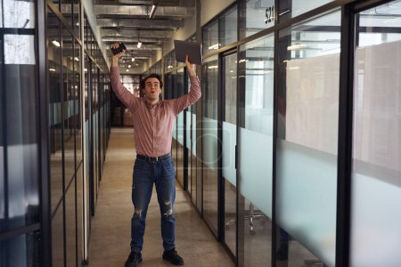 Photo for Full-size portrait of jubilant young corporate worker with laptop and smartphone in raised hands looking up at ceiling in hallway - Royalty Free Image