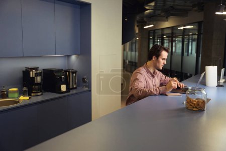 Photo for Focused company employee with cookie in hand sitting at table while looking at laptop screen at snack time - Royalty Free Image