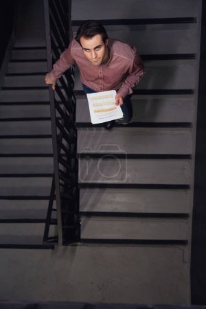 Téléchargez les photos : Vue de dessus du jeune employé d'entreprise avec la documentation d'affaires descendant les escaliers et regardant vers le haut - en image libre de droit