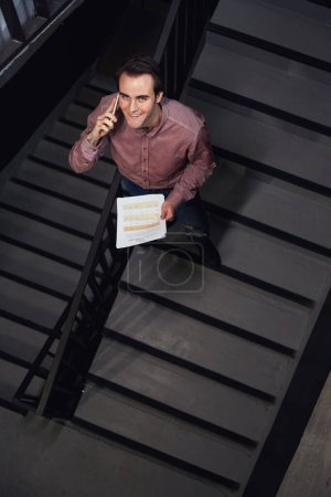Téléchargez les photos : Vue du dessus du joyeux employé de bureau avec pile de documents parlant sur smartphone sur escalier et regardant vers le haut - en image libre de droit