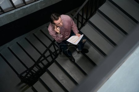 Photo for Top view of corporate worker with pile of documents standing on stairs during smartphone conversation - Royalty Free Image