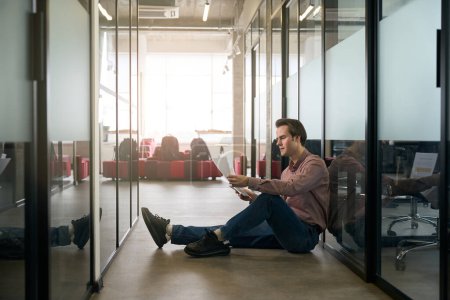 Téléchargez les photos : Employé de l'entreprise ciblée avec smartphone et papiers dans les mains assis sur le sol par un mur de verre dans le couloir - en image libre de droit