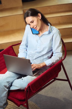 Photo for Smiling young woman seated in armchair typing on laptop keyboard while talking on mobile phone - Royalty Free Image
