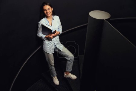 Téléchargez les photos : Portrait grandeur nature de dame gaie avec téléphone portable et ordinateur portable dans les mains debout sur un escalier en colimaçon - en image libre de droit