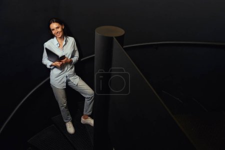 Téléchargez les photos : Portrait complet de femme joyeuse tenant smartphone et ordinateur portable dans les mains tout en se tenant debout sur les escaliers dans le bâtiment - en image libre de droit