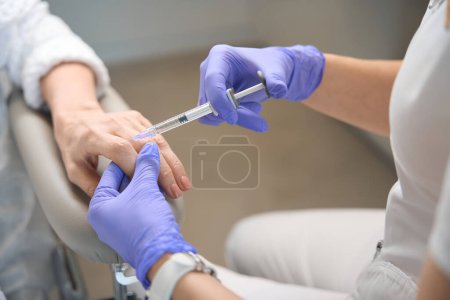 Photo for Close up of cosmetologist hands in gloves performing injection on mature lady knuckle at beautician office - Royalty Free Image
