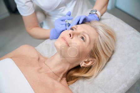 Téléchargez les photos : Mains de cosmétologue effectuant l'injection sur pommette dame mature dans le salon de beauté - en image libre de droit