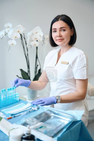 Téléchargez les photos : Dermatologue en gants assis au bureau de cosmétologie devant des affaires médicales sur le bureau - en image libre de droit