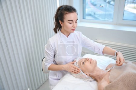 Téléchargez les photos : Cosmetologue effectuant une procédure de beauté sur le cou pour femme mature couchée sur le canapé dans le bureau de l'esthéticienne - en image libre de droit