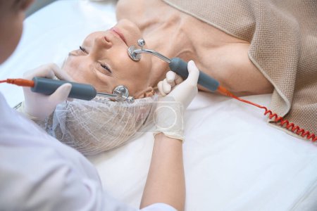 Photo for Cosmetician doing microcurrent therapy on mature womans face in modern clinic - Royalty Free Image