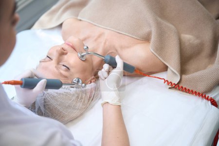 Photo for Top view of cosmetician doing microcurrent therapy on mature woman face in modern clinic - Royalty Free Image