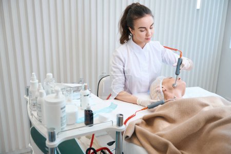 Téléchargez les photos : Cosméticien médical faisant une thérapie à microcourant sur le visage féminin mature dans une clinique moderne - en image libre de droit