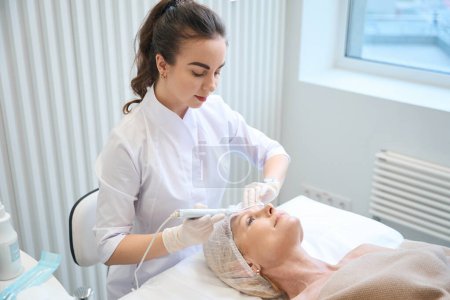 Téléchargez les photos : Esthéticienne médicale faisant nettoyage cosmétique du visage pour femme adulte dans un salon de beauté - en image libre de droit