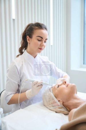 Photo for Smiling beautician making cosmetic facial cleansing for adult lady in beauty salon - Royalty Free Image