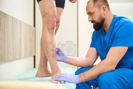 Téléchargez les photos : Homme médecin dans un masque et des gants médicaux effectue un examen des jambes d'un patient - en image libre de droit