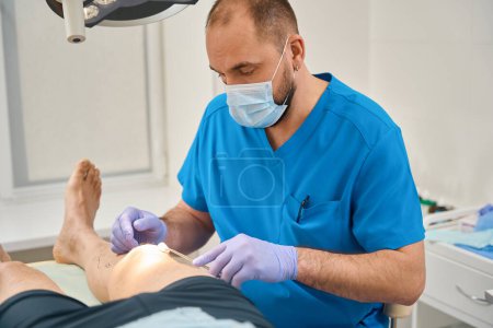 Photo for Male doctor in operating room swabs the leg of a patient lying on operating table - Royalty Free Image
