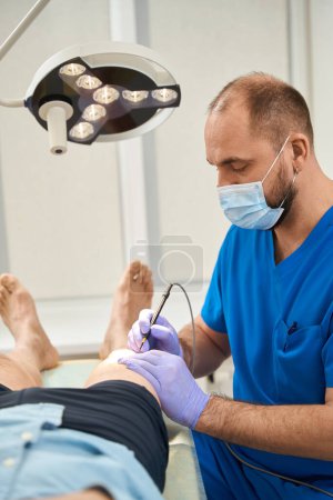 Photo for Vertical photo of a male surgeon in a blue coat during surgery on the leg of an elderly male patient - Royalty Free Image