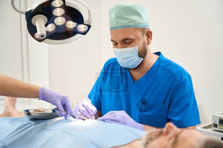 Foto de Cirujano con abrigo azul durante una operación en el cuerpo de un anciano acostado en una mesa - Imagen libre de derechos