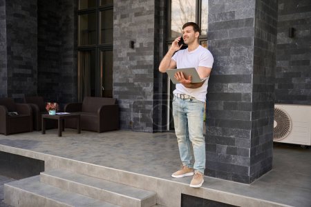 Téléchargez les photos : Homme dans un T-shirt blanc se tient près de l'entrée de la maison avec des ordinateurs portables - en image libre de droit