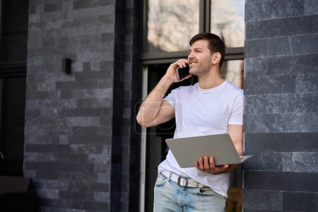 Photo for Smiling guy talking on the phone with a laptop in one hand - Royalty Free Image