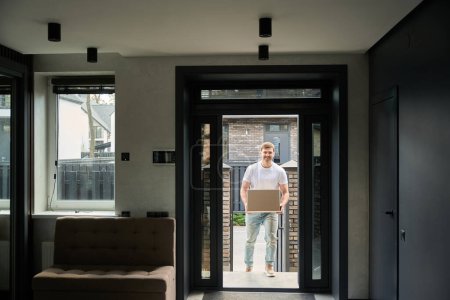 Photo for Young guy in a white T-shirt brings a box into his new home - Royalty Free Image